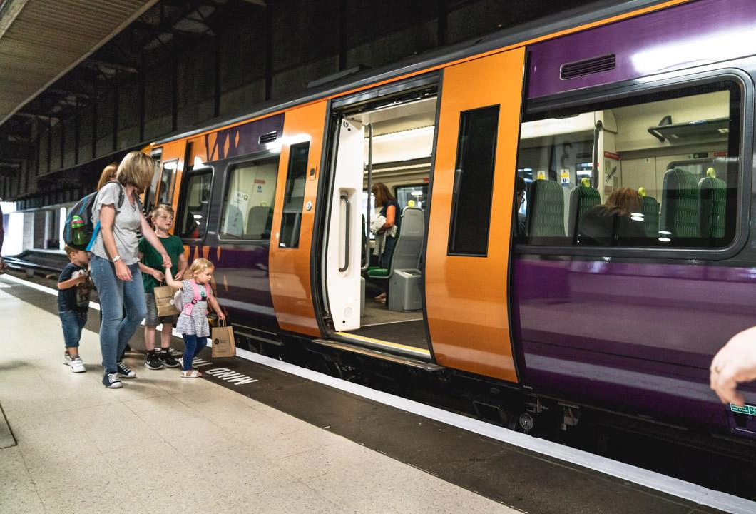 A purple and yellow West Midlands Railway train sat stationary at a platform at a train station, with a set of doors open and a group of adults and children about to board.