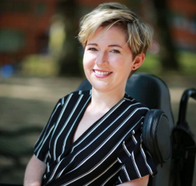 Sarah Rennie, a white woman with short blonde hair sitting in her powerchair smiling towards the camera