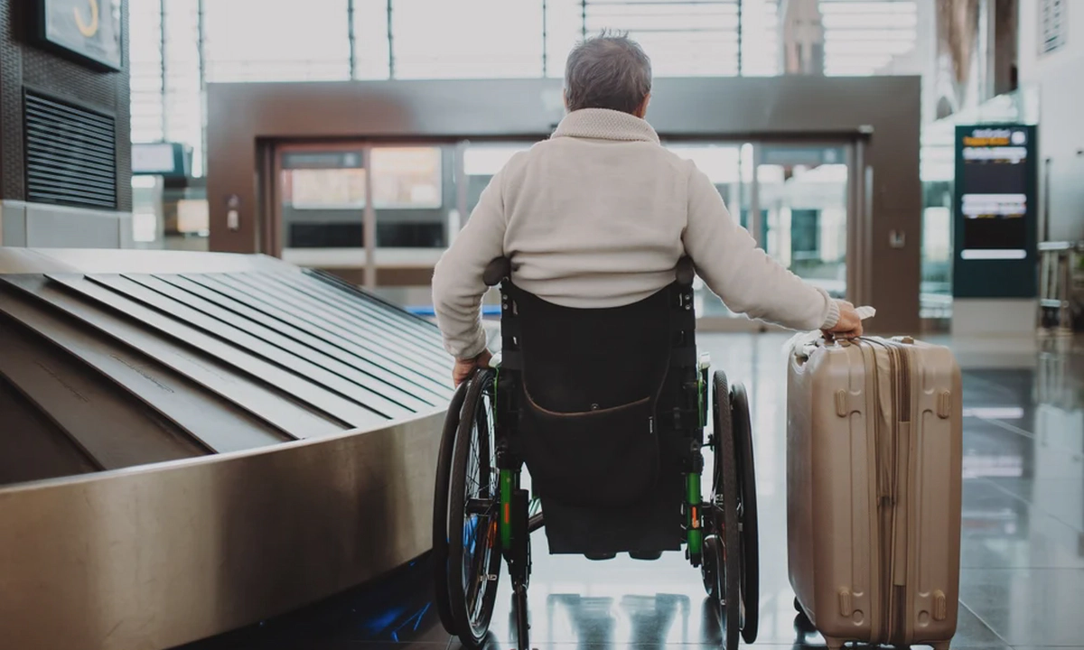 A man wearing a white jumper sitting in a wheelchair with his back to the camera, holding a suitcase on wheels in his right hand, in an airport. A man wearing a white jumper sitting in a wheelchair with his back to the camera, holding a suitcase on wheels in his right hand, in an airport.