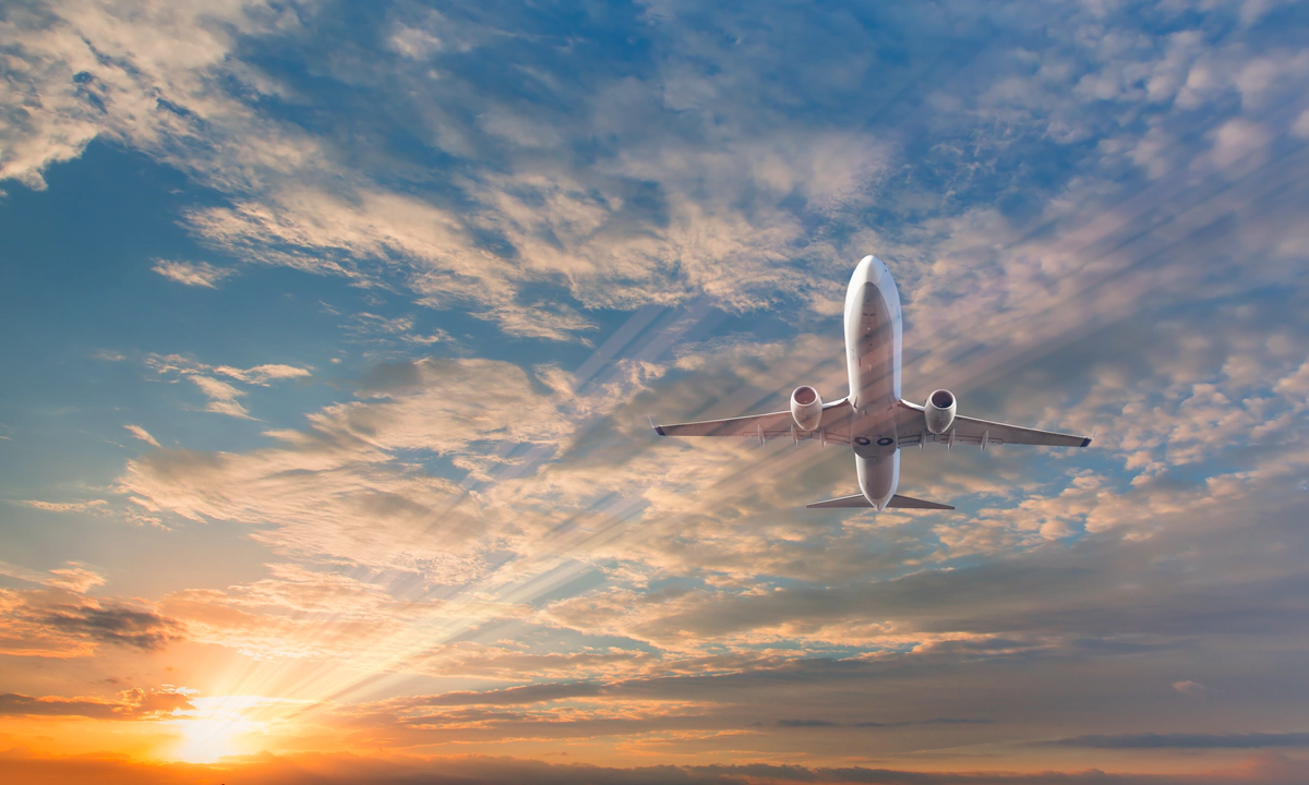 A picture of a white passenger plane from below, against a blue sky filled with clouds and a lens flare from the sun A picture of a white passenger plane from below, against a blue sky filled with clouds and a lens flare from the sun