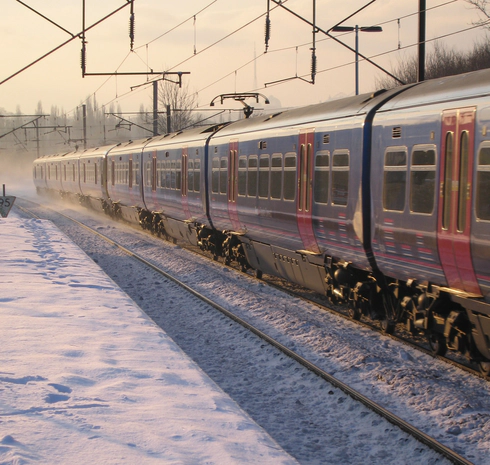Photo of a blue and red train on a snowy track beside a snowy platform with an orange sky behind it.