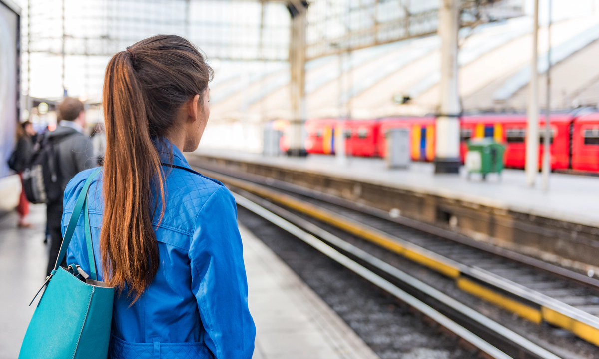 Photo of a train platform. The platform and people on the platform are blurred except for a woman in the foreground who stands with her back to the camera with long brown hair in a ponytail and a blue denim jacket. A red train is waiting at the platform opposite. Photo of a train platform. The platform and people on the platform are blurred except for a woman in the foreground who stands with her back to the camera with long brown hair in a ponytail and a blue denim jacket. A red train is waiting at the platform opposite.