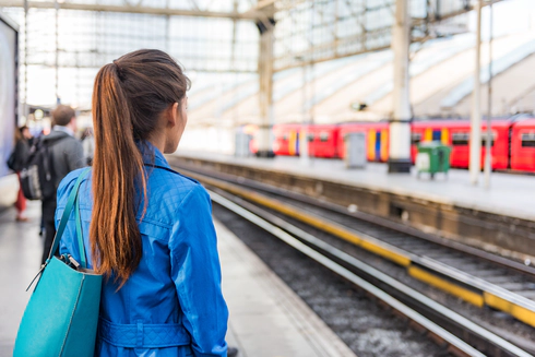 Photo of a train platform. The platform and people on the platform are blurred except for a woman in the foreground who stands with her back to the camera with long brown hair in a ponytail and a blue denim jacket. A red train is waiting at the platform opposite.