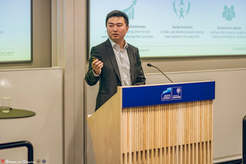 Jay Shen, an asian man with short black hair, and CEO of Transreport, in a grey suit standing at a speaking podium.