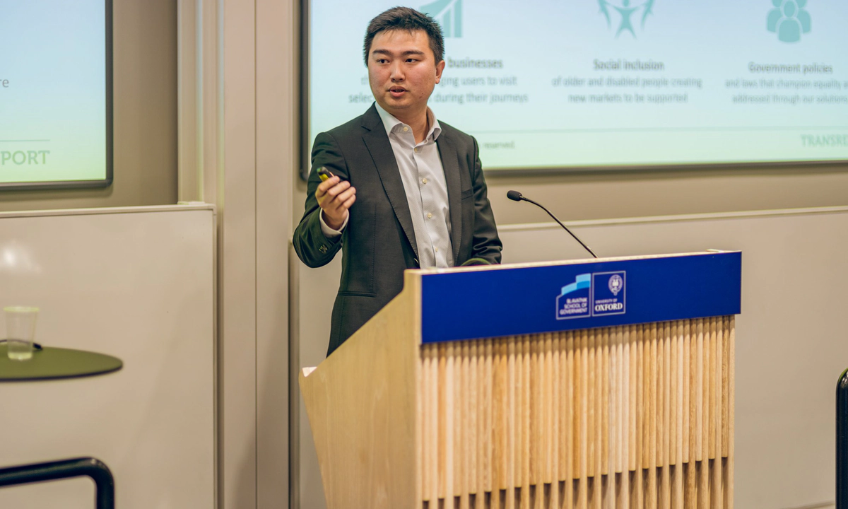 Jay Shen, an asian man with short black hair, and CEO of Transreport, in a grey suit standing at a speaking podium. Jay Shen, an asian man with short black hair, and CEO of Transreport, in a grey suit standing at a speaking podium.