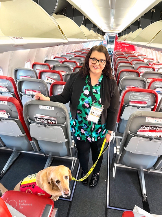 Emma Partlow, Transreport's Inclusion & Accessibility Manager, standing beside her assistance dog Luna, in the middle aisle of a commercial passenger aeroplane.
