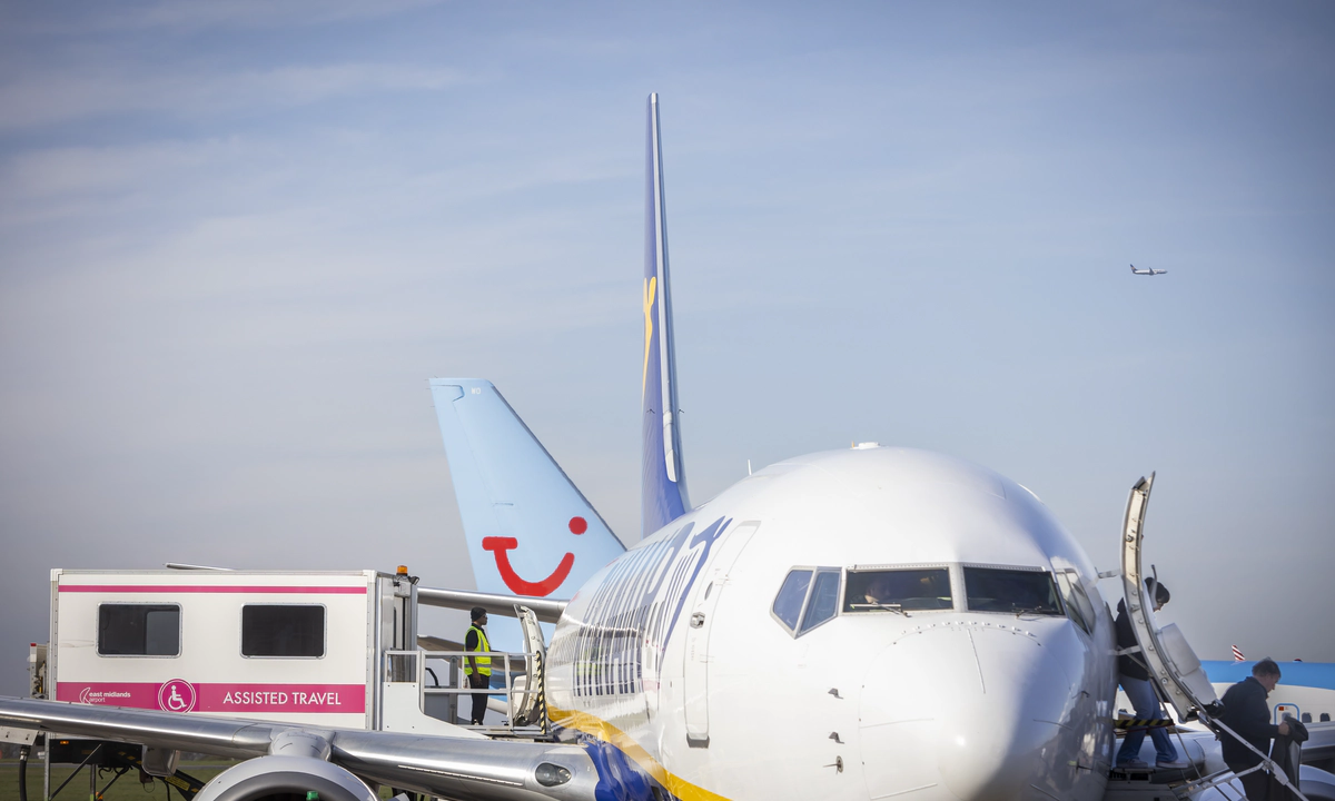 A zoomed in photograph of a Ryainair Boeing 747 plane from the front, with passengers disembarking by the front exit, by stairs, on the right, and an ambulift docking with the central exit of the plane on the left. A zoomed in photograph of a Ryainair Boeing 747 plane from the front, with passengers disembarking by the front exit, by stairs, on the right, and an ambulift docking with the central exit of the plane on the left.